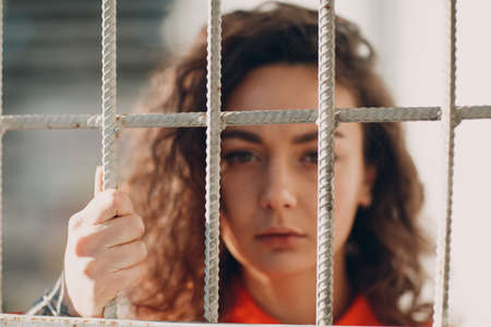 Young brunette curly woman in orange suit behind jail bars. Female in colorful overalls portraitの写真素材