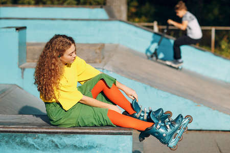 Young redhaired woman in green and yellow clothes and orange stockings with curly hairstyle put on rollerblades in skate roller parkの写真素材