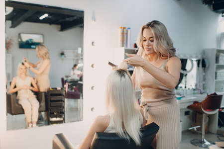 Hairdresser female making hair extensions to young woman with blonde hair in beauty salon. Professional hair extensionの写真素材