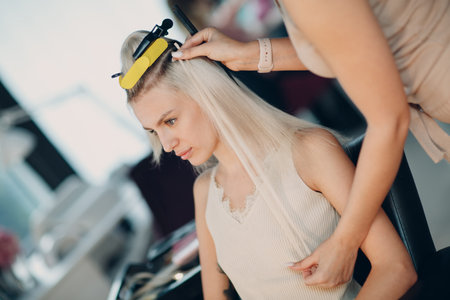 Hairdresser female making hair extensions to young woman with blonde hair in beauty salon. Professional strand hair extensionの写真素材