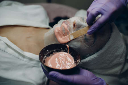 Alginate face mask application process. A female hands of a beautician holding spatula. Woman lying on a couch in office of cosmetologist. Facial skincare in the beauty spa salon.の写真素材