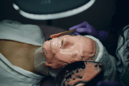 Alginate face mask application process. A female hands of a beautician holding spatula. Woman lying on a couch in office of cosmetologist. Facial skincare in the beauty spa salon.の写真素材
