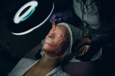 Alginate face mask application process. A female hands of a beautician holding spatula. Woman lying on a couch in office of cosmetologist. Facial skincare in the beauty spa salon.の写真素材