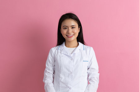 Asian positive doctor woman in white medical white gown against pink background portraitの写真素材