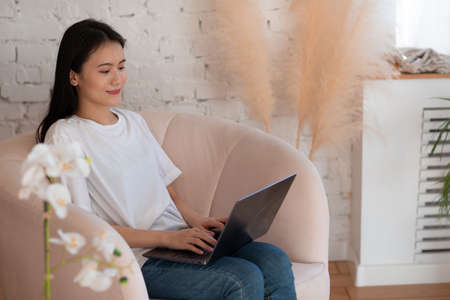 Portrait of asian woman enjoying using laptop at apartment interior indoor living room. Video call vlogger webinar web stream conceptの写真素材