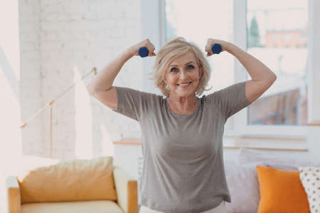 Elderly caucasian old aged woman portrait gray haired showing biceps muscles doing exercises with dumbbells in casual wear at home apartment living roomの写真素材