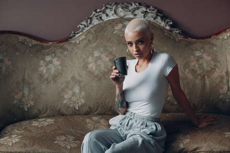 Millenial young european woman with short blonde hair portrait sitting with cup of tea or coffee drink on vintage sofaの写真素材
