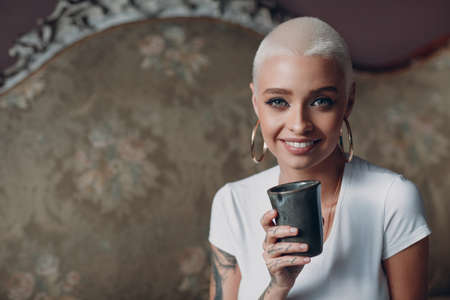 Millenial young european english woman with short blonde hair portrait sitting with cup of tea or coffee drink on vintage sofaの写真素材