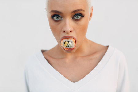 Portrait of young smiling millenial european short haired woman with sushi Philadelphia salmon roll in her mouth . Beautiful happy blonde girl outdoor.の写真素材