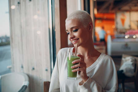 Portrait of young smiling millenial european short haired woman with green smoothie at cafe. Beautiful happy blonde girl indoor.の写真素材