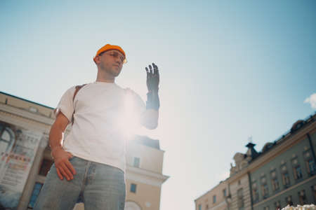 Young disabled man with artificial prosthetic hand in casual clothes at city street looking to his hand against blue skyの写真素材