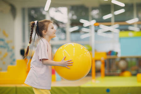 Little girl playing and doing exercises with big yellow ball in gym at kindergarten or elementary...