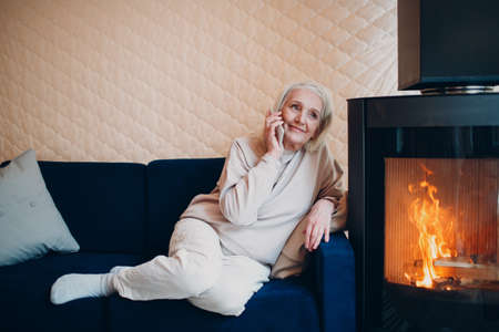 Elderly gray haired woman sitting on sofa and talking smartphone in living room with fireplace chimney.の写真素材
