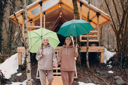Elderly and young adult women with umbrella at glamping camping tent. Modern autumn vacation lifestyle conceptの写真素材