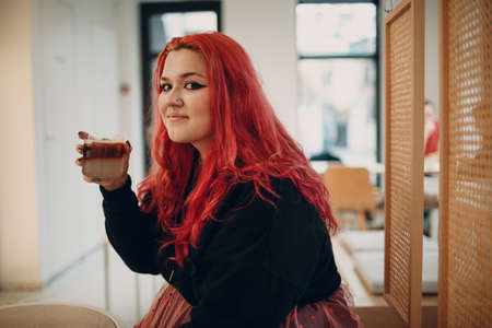 European plus size woman. Young red pink haired body positive girl sitting in cafe with coffee drinkの写真素材