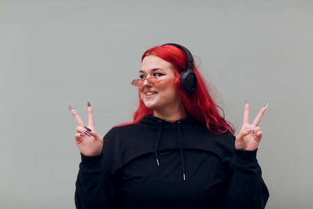 European plus size woman in headphones showing V victory fingers sign. Young red pink haired body...