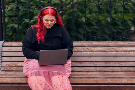 European plus size woman uses laptop while sitting on wooden bench outdoors at daytime. Young focused red pink haired girl wears glasses and headphones.の写真素材