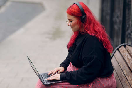 European plus size woman uses laptop while sitting on wooden bench outdoors at daytime. Young focused red pink haired body positive girl wears glasses and headphones.の写真素材