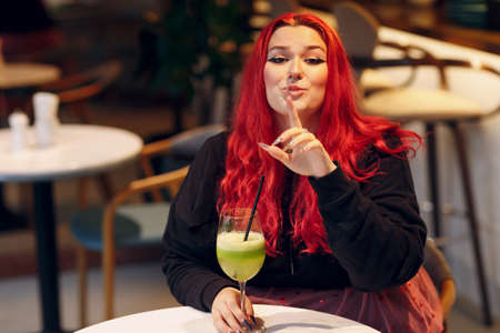 European plus size woman drink green smoothie in cafe. Young red pink haired body positive girl sitting at restaurantの写真素材