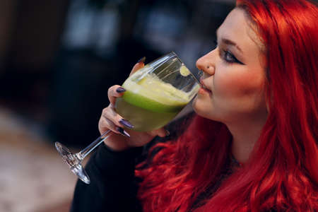 European plus size woman drink green smoothie in cafe. Young red pink haired body positive girl sitting at restaurantの写真素材