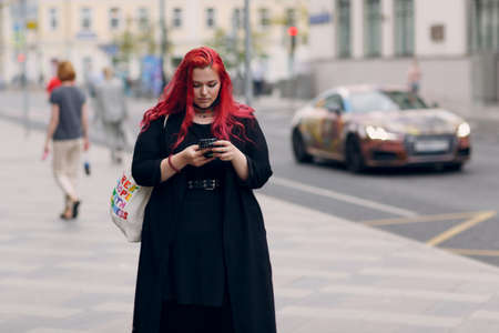 European plus size woman walking outdoor street city with mobile cellular phone. Young red pink haired body positive girl using cellphone smartphoneの写真素材
