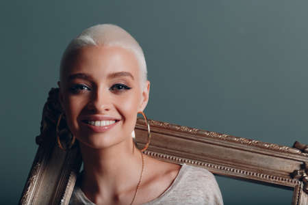 Millenial young woman with short blonde hair holds gilded picture frame in hands on her neck...