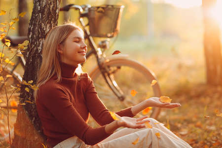 Happy young woman sitting near vintage bicycle in autumn park at sunset. Fall season concept.の写真素材