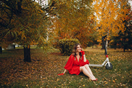 Young woman with electric scooter in red dress at the autumn fall city park.の写真素材