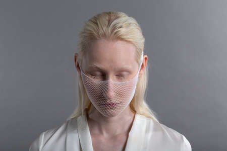 Closeup portrait of white caucasian albino blond woman fashion model wearing quarantine medical face mask coarse mesh net on grey background. face coveringの写真素材