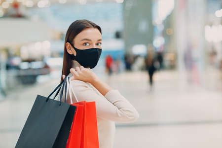 Young adult woman in protective medical face mask carrying paper shopping bags in hands at supermarket.の写真素材