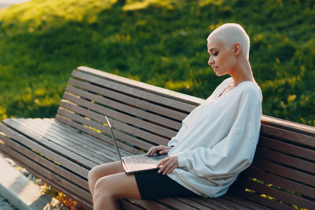 Portrait of young smiling millenial european short haired woman using laptop at bench on green grass meadow in park. Beautiful happy blonde girl outdoor.の写真素材