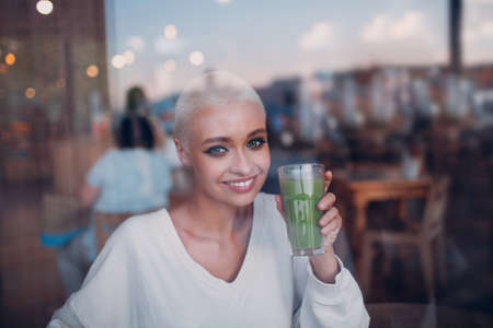 Portrait of young smiling millenial european short haired woman with green smoothie behind window glass at cafe. Beautiful happy blonde girl indoor.の写真素材