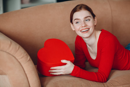 Beautifil young woman with red hair lying in her sofa in living room and holding giving gift box in the shape of heart and smile. Valentines Dayの写真素材