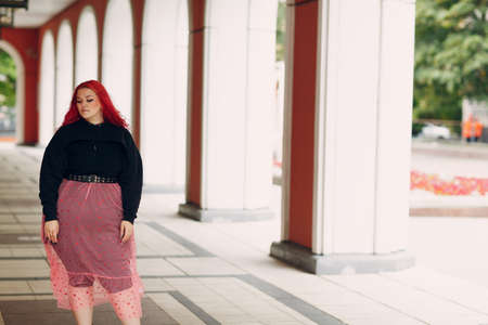 European plus size bold woman walking outdoor street city. Young red pink haired body positive girl.の写真素材
