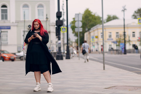 European bold plus size woman walking outdoor street city with mobile cellular phone. Young red pink haired body positive girl using cellphone smartphoneの写真素材