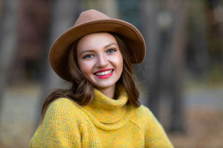 Portrait of young smiling woman model in hat and yellow sweater at autumn park with yellow foliage leaves. Fall season fashion concept.の写真素材