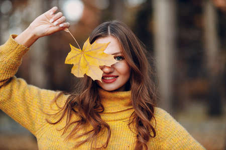 Young woman model in autumn park with yellow foliage maple leaf at face. Fall season fashionの写真素材