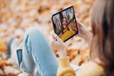 Young woman model take selfie photo with tablet pc in autumn park with yellow foliage maple leaves. Fall season fashionの写真素材
