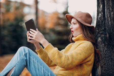 Young woman model surprised with tablet pc in autumn park with yellow foliage maple leaves. Online shopping concept.の写真素材