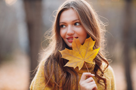 Young woman model in autumn park with yellow foliage maple leaf at face. Fall season fashionの写真素材