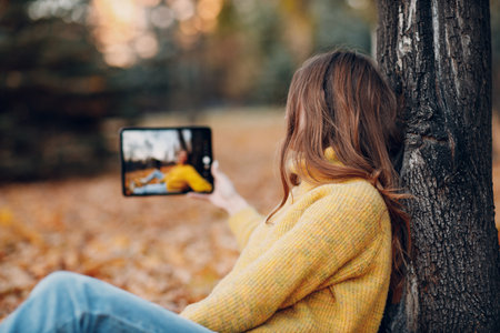 Young woman model take selfie photo with tablet pc in autumn park with yellow foliage maple leaves. Fall season fashionの写真素材