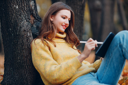 Young woman model sitting with tablet pc and painting with pencil stylus in autumn park with yellow foliage maple leaves.の写真素材