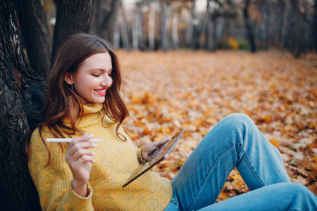 Young woman model sitting with tablet pc and painting with pencil stylus in autumn park with yellow foliage maple leaves.の写真素材