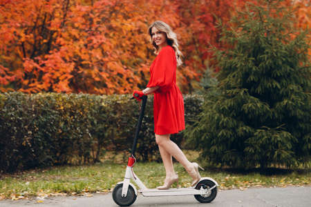 Young woman in red dress ride electric scooter in autumn city. Alternative commuter transportation.の写真素材