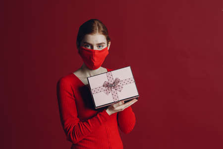 Young woman holding christmas gift box present in face mask. Cute surprised girl on red background.の写真素材