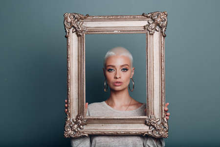 Millenial young woman with short blonde hair holds gilded picture frame in hands behind her face portraitの写真素材
