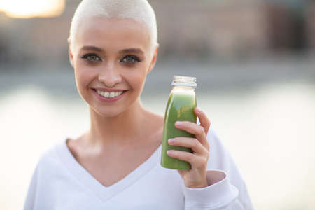 Portrait of young smiling millenial european short haired woman with green smoothie. Beautiful happy blonde girl outdoor.の写真素材