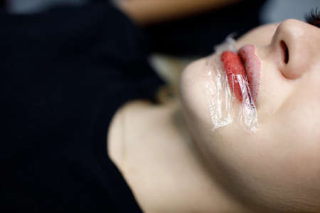Cosmetologist applying red permanent make up tattoo on young woman lips. Permanent lips tattoo procedure cellophane polyethyleneの写真素材