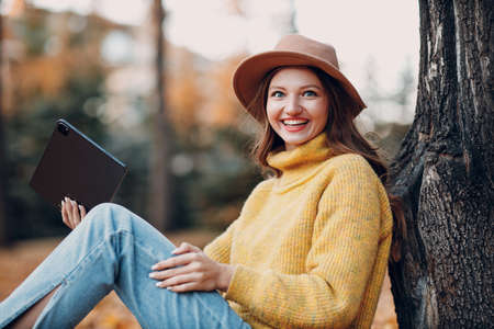 Young woman model sitting with tablet pc in autumn park with yellow foliage maple leaves. Fall season fashionの写真素材