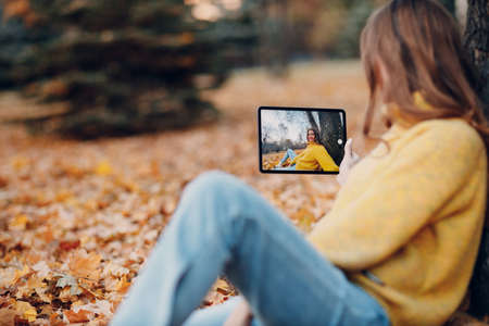 Young woman model take selfie photo with tablet pc in autumn park with yellow foliage maple leaves. Fall season fashionの写真素材